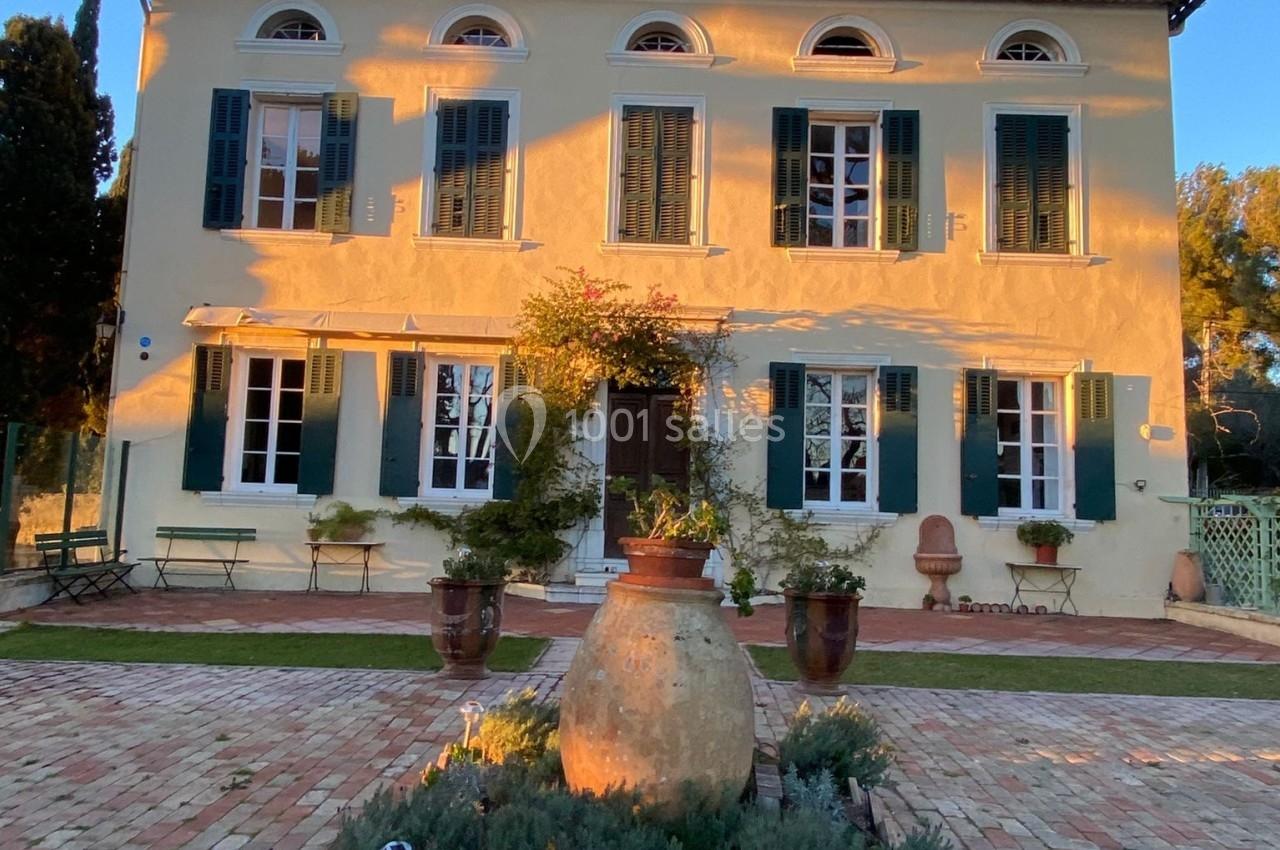 Façade d'une maison provençale jaune à volets verts, éclairée par le soleil couchant, avec une cour pavée et des plantes.