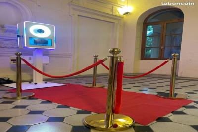 Miniature Traiteur Lattes (Hérault) - A la Bonne Franquette #41 Borne photo interactive éclairée entourée de poteaux dorés et cordons rouges sur un tapis rouge dans une salle lumineuse.