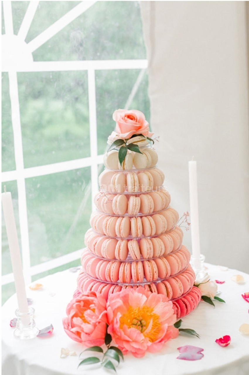 Tour de macarons roses et blancs décorée de fleurs, placée sur une table près d'une fenêtre lumineuse.