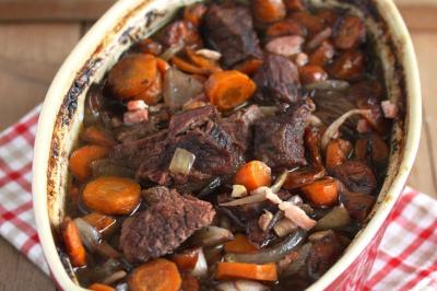 Plat de tajine en terre cuite contenant un mélange de légumes, olives et viande, avec théière et couscous en arrière-plan.
