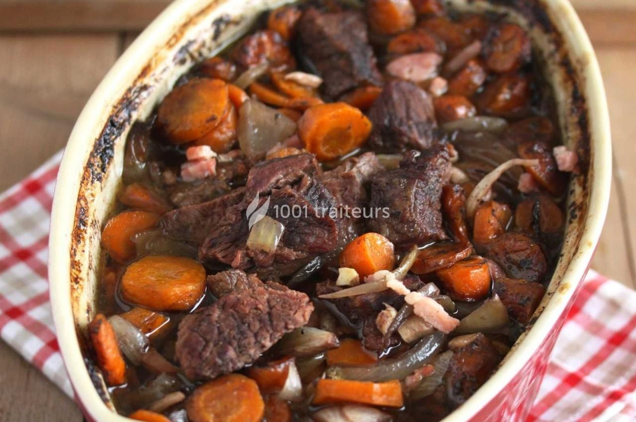Plat mijoté avec morceaux de viande, carottes, oignons et lardons dans un plat en céramique sur une nappe à carreaux.