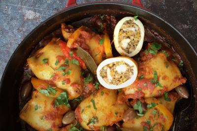 Plat de tajine en terre cuite contenant un mélange de légumes, olives et viande, avec théière et couscous en arrière-plan.