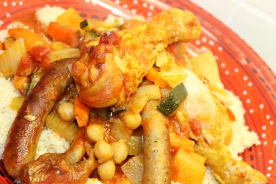 Plat de tajine en terre cuite contenant un mélange de légumes, olives et viande, avec théière et couscous en arrière-plan.