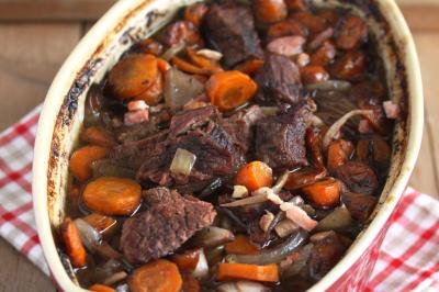 Plat de tajine en terre cuite contenant un mélange de légumes, olives et viande, avec théière et couscous en arrière-plan.