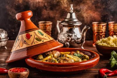 Plat de tajine en terre cuite contenant un mélange de légumes, olives et viande, avec théière et couscous en arrière-plan.