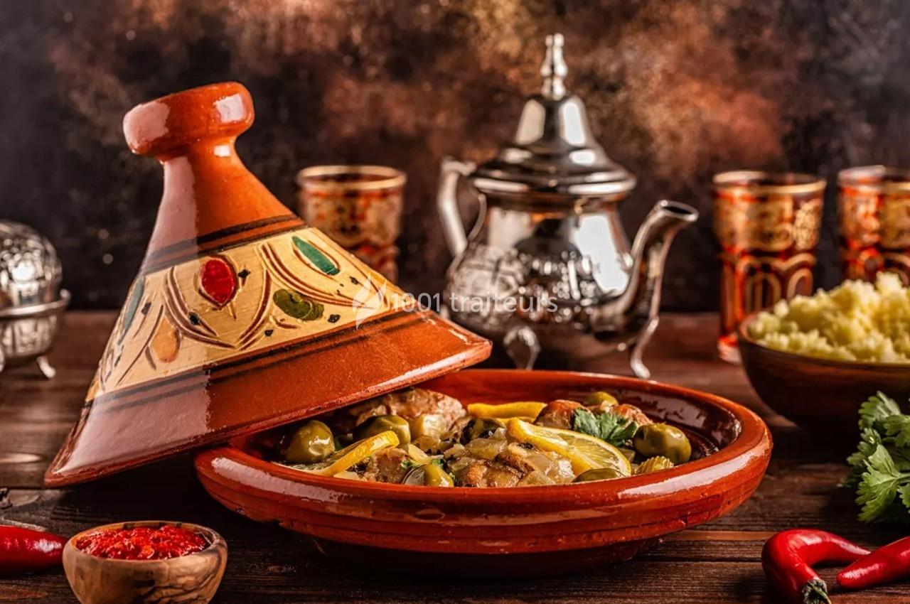 Plat de tajine en terre cuite contenant un mélange de légumes, olives et viande, avec théière et couscous en arrière-plan.
