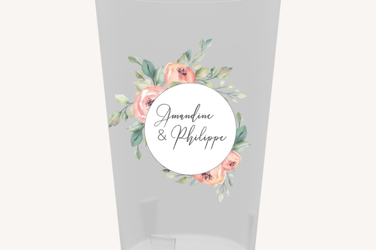 Verre transparent décoré d'une étiquette ronde avec des fleurs pastel et des prénoms manuscrits au centre.