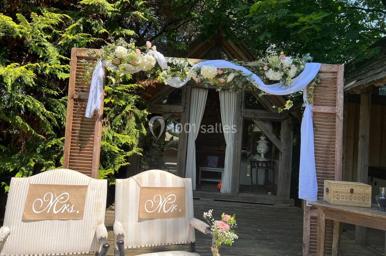 Arche de mariage décorée de fleurs et de voilages blancs, avec deux fauteuils ’Mrs.’ et ’Mr.’ devant un bâtiment en bois.