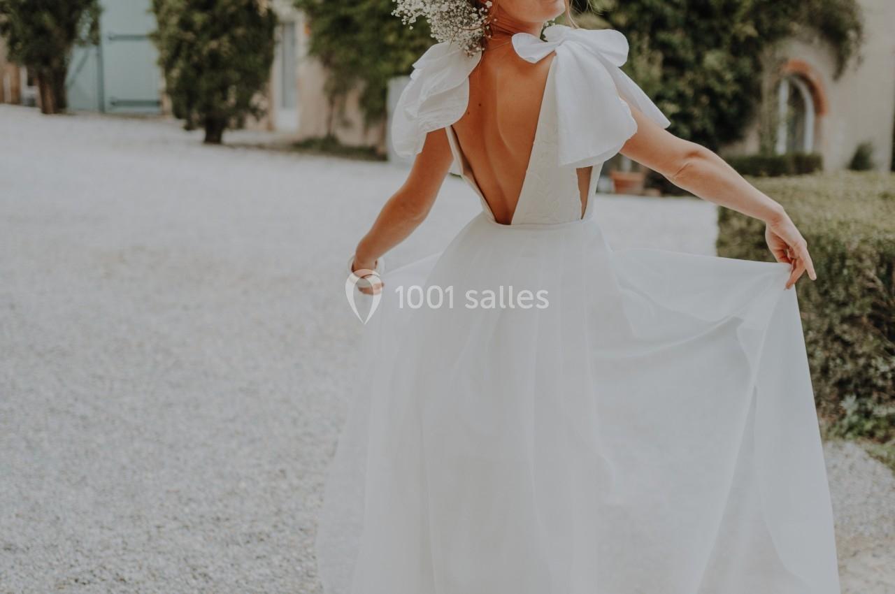 Femme en robe de mariée dos nu blanche, tenant le tissu fluide, devant un bâtiment entouré de verdure.