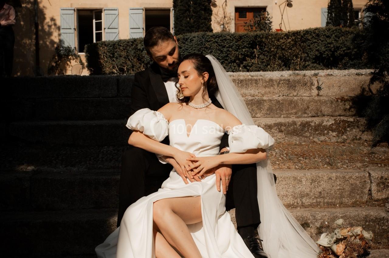Un couple de mariés assis sur des marches en pierre devant une maison aux volets bleus, dans une lumière douce.