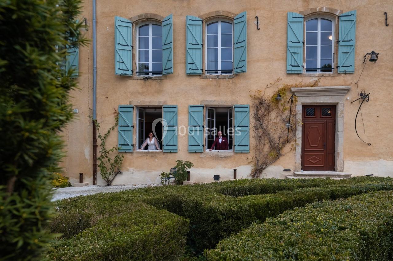 Façade d'une maison ancienne avec volets bleus, deux personnes regardant par des fenêtres ouvertes, jardin au premier plan.
