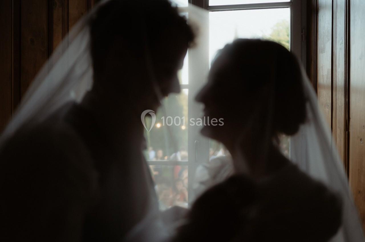 Deux silhouettes floues d'un couple sous un voile, devant une fenêtre laissant passer la lumière naturelle.