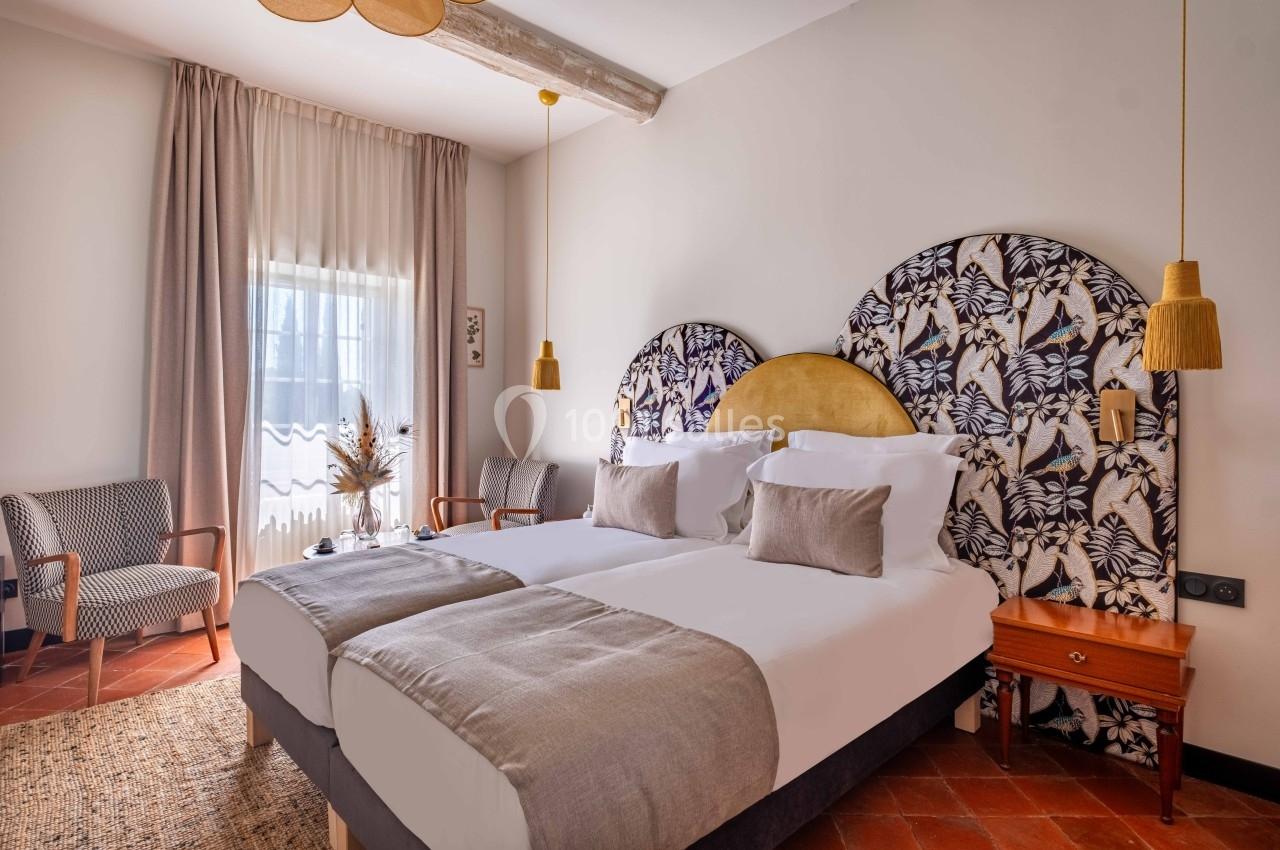 Chambre lumineuse avec deux lits simples, tête de lit décorative, lampes suspendues et fauteuils près d'une fenêtre.