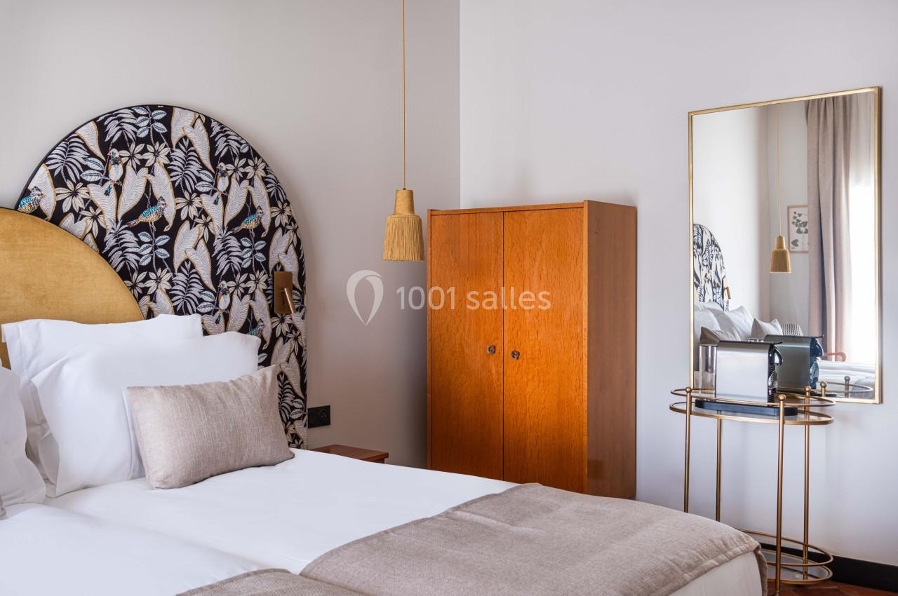 Chambre lumineuse avec lit double, tête de lit décorative, armoire en bois, miroir et petite table dorée.