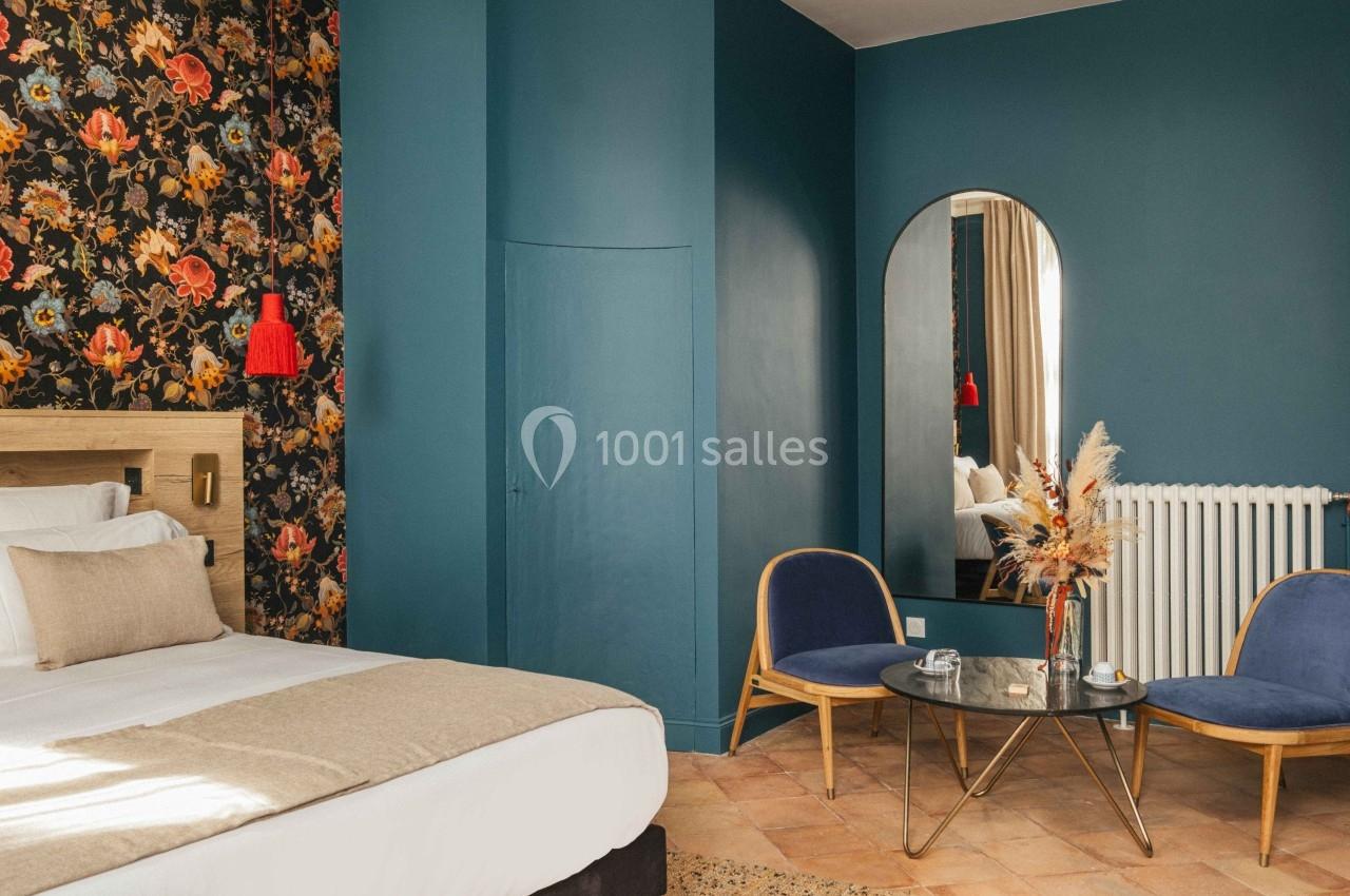 Chambre décorée avec un mur bleu, papier peint fleuri, lit double, deux fauteuils bleus et une table basse.