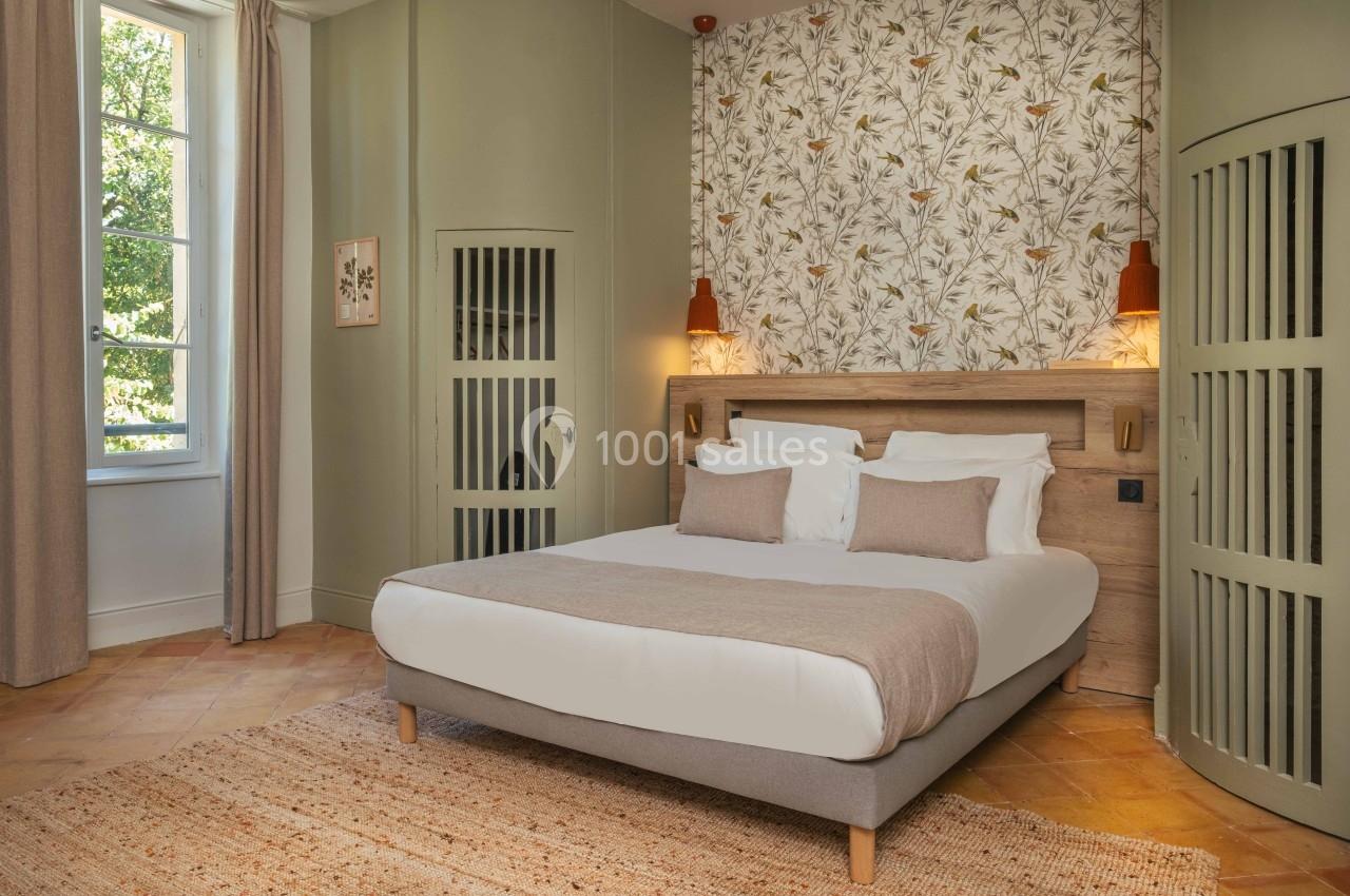 Chambre lumineuse avec un lit double, mur décoré de papier peint floral, éclairage mural et fenêtre donnant sur un jardin.