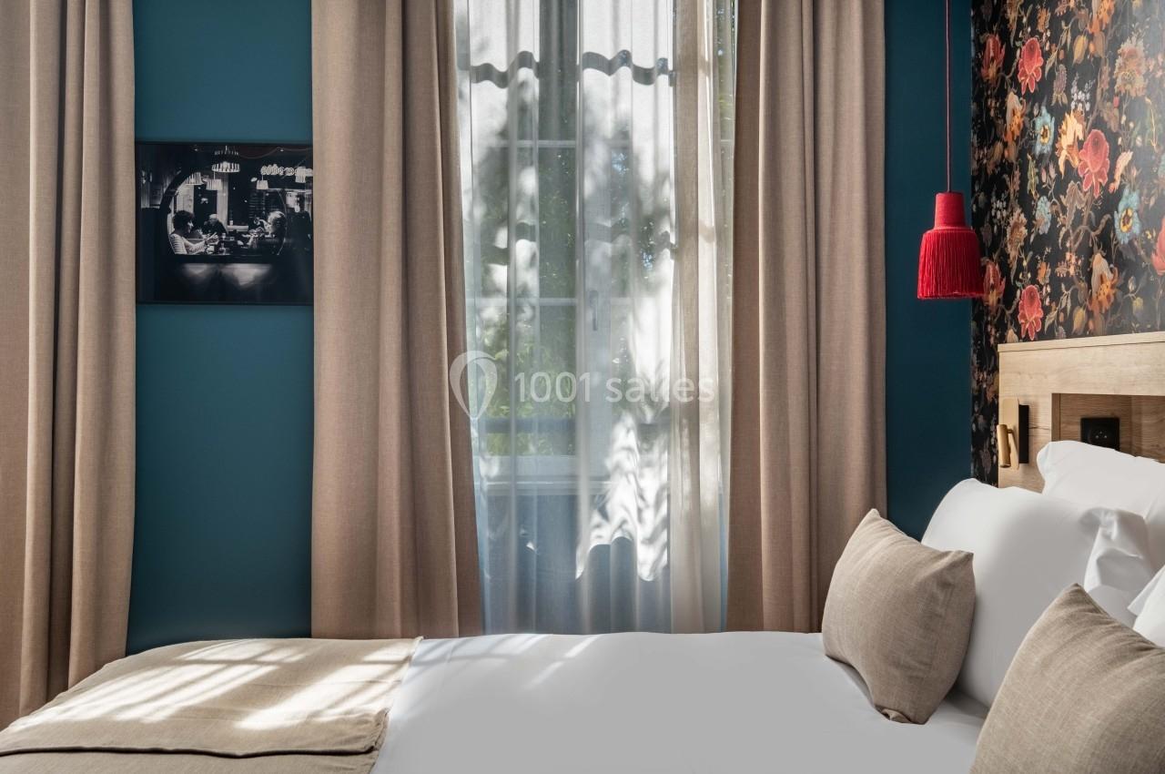 Chambre lumineuse avec lit double, rideaux beiges, mur bleu, décoration florale et lampe rouge suspendue.