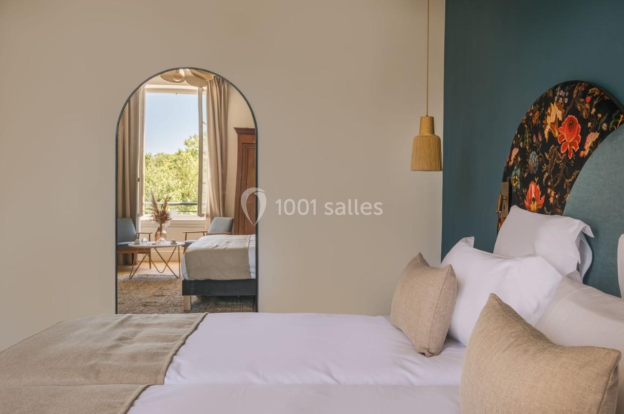 Chambre lumineuse avec lit double, tête de lit fleurie, miroir en arche reflétant un coin salon et une fenêtre ouverte.