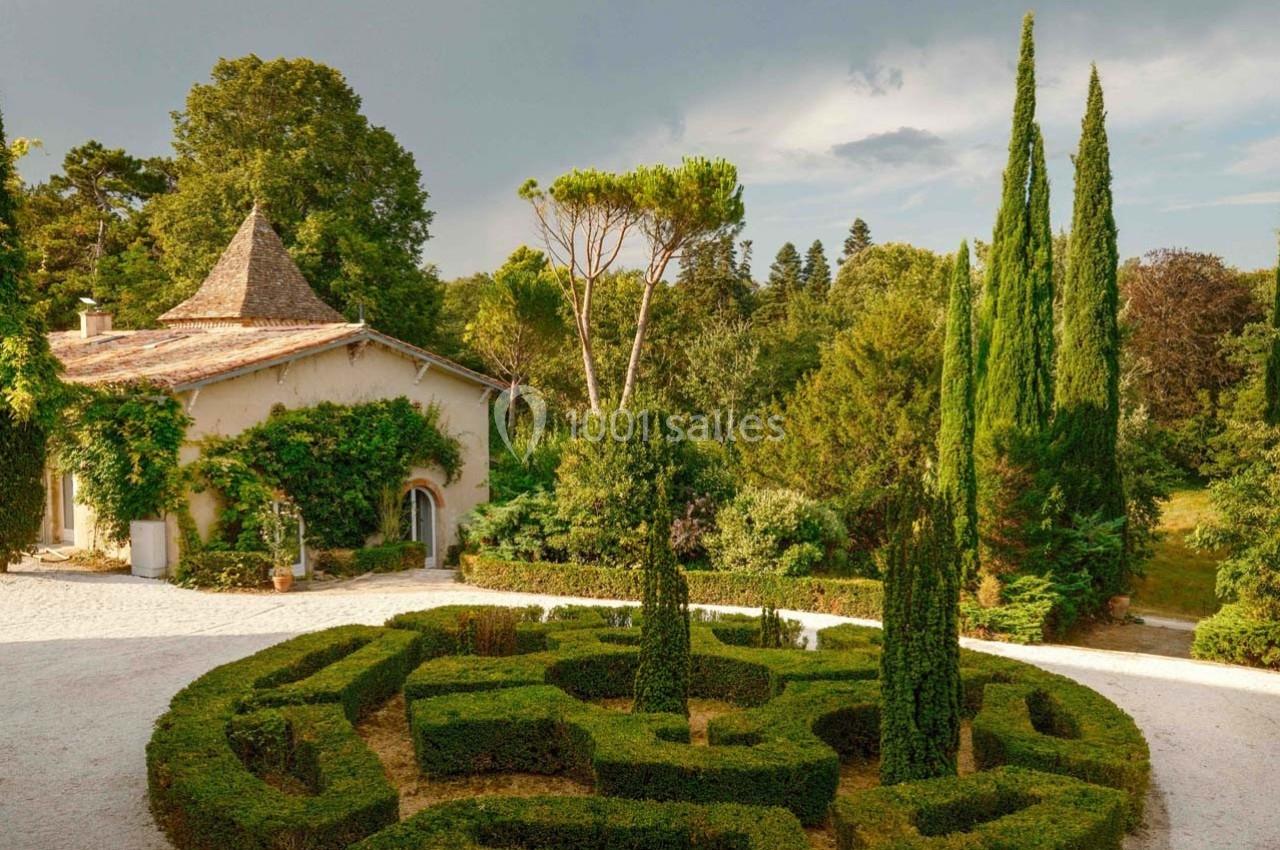 Jardin avec haies taillées en labyrinthe devant une maison en pierre entourée de végétation et de grands arbres.
