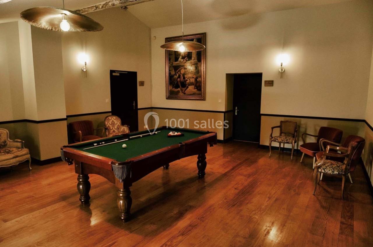 Salle avec un billard au centre, entourée de chaises, éclairée par des lampes suspendues et un tableau mural.