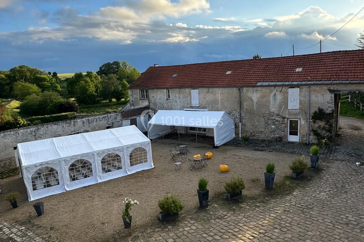 Cour pavée avec tentes blanches installées près d'un bâtiment ancien, entourée de pots de plantes et d'un paysage rural.