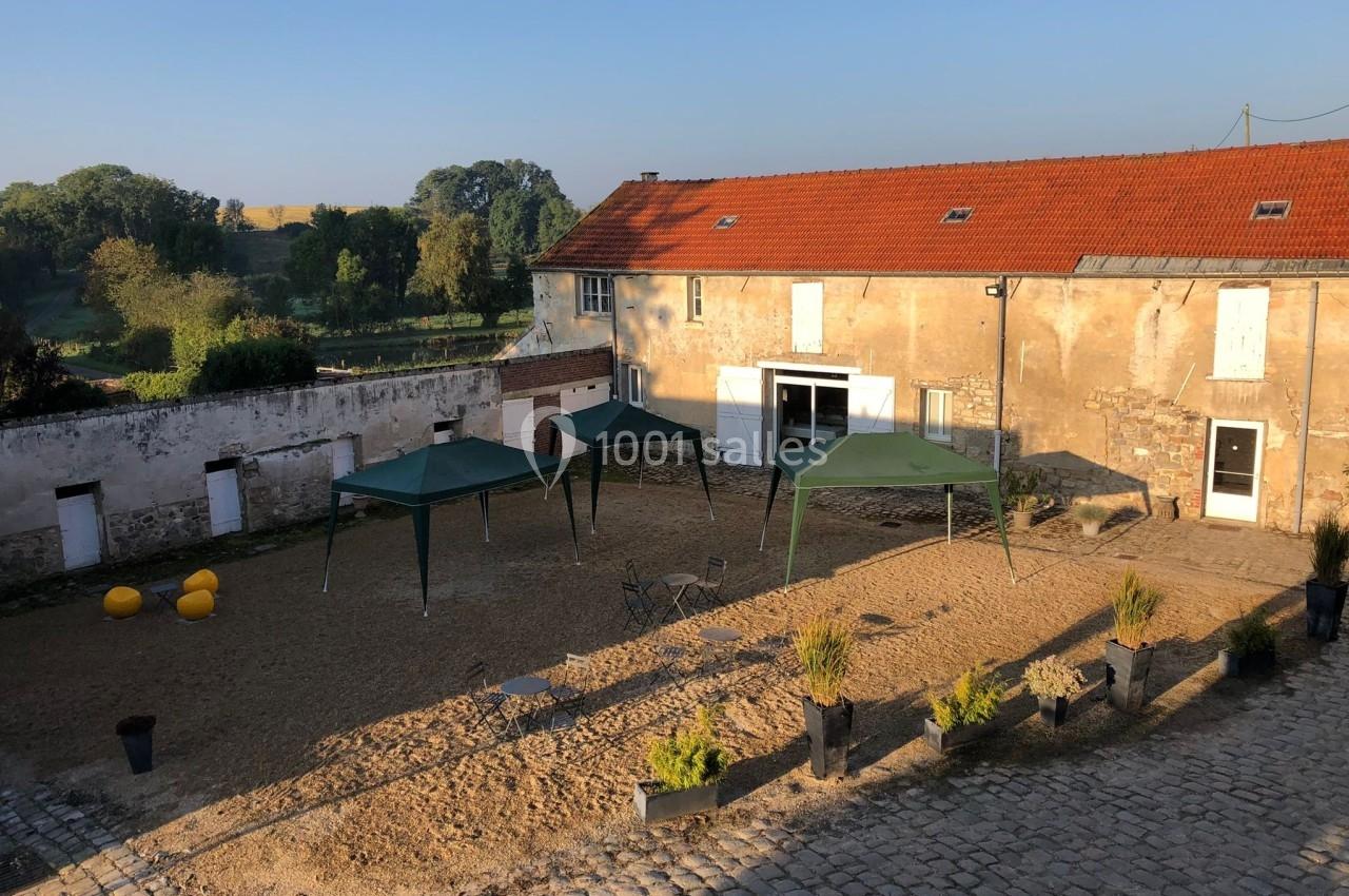 Cour ensoleillée avec sol en sable, trois tonnelles vertes, quelques plantes en pot et un bâtiment en pierre aux toits…