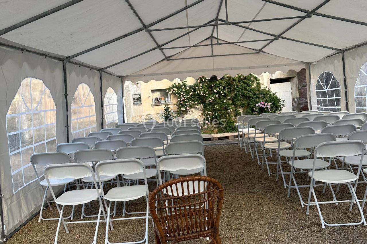 Chaises blanches alignées sous une tente blanche ouverte, avec une chaise en osier au premier plan.