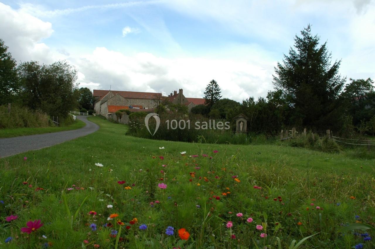 Chemin bordé de fleurs sauvages menant à des bâtiments en pierre entourés de verdure sous un ciel nuageux.