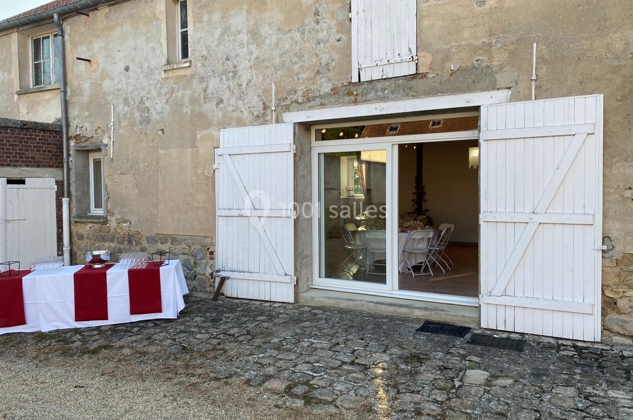 Façade d'un bâtiment ancien avec porte vitrée ouverte sur une salle aménagée, table dressée à l'extérieur.
