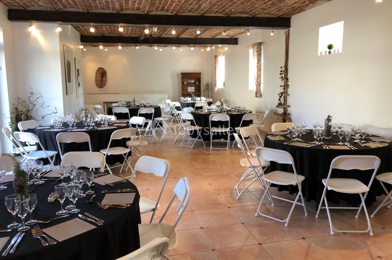 Salle lumineuse avec tables rondes dressées de nappes noires, chaises blanches pliantes et décorations sobres.