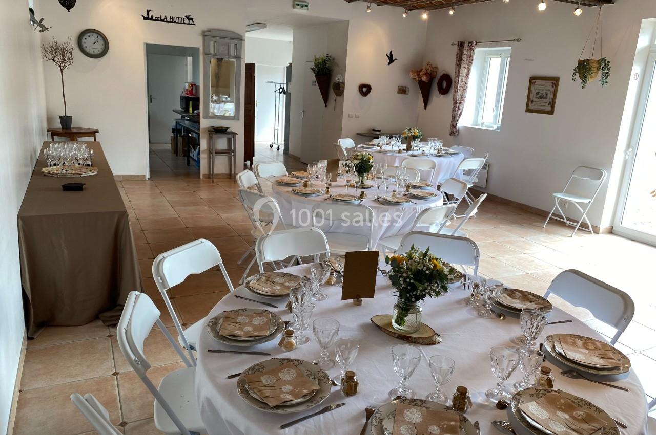Salle lumineuse décorée pour un repas, avec tables rondes dressées, nappes blanches et chaises pliantes blanches.