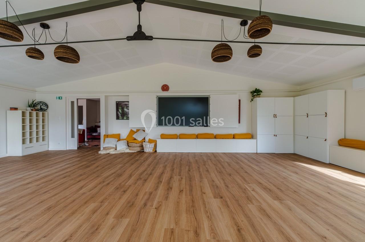 Salle lumineuse avec parquet, coussins jaunes, écran mural, rangements blancs et suspensions décoratives.