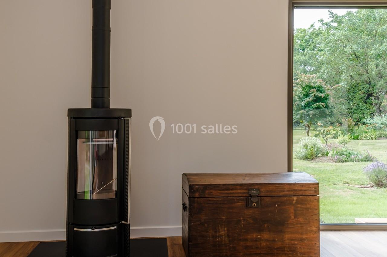 Poêle à bois noir sur un sol en parquet, à côté d'un coffre en bois, avec vue sur un jardin par une baie vitrée.