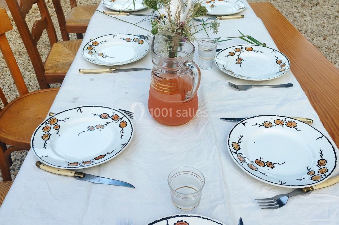 Table extérieure dressée avec une nappe blanche, des assiettes décorées, des couverts et une carafe de boisson orange.