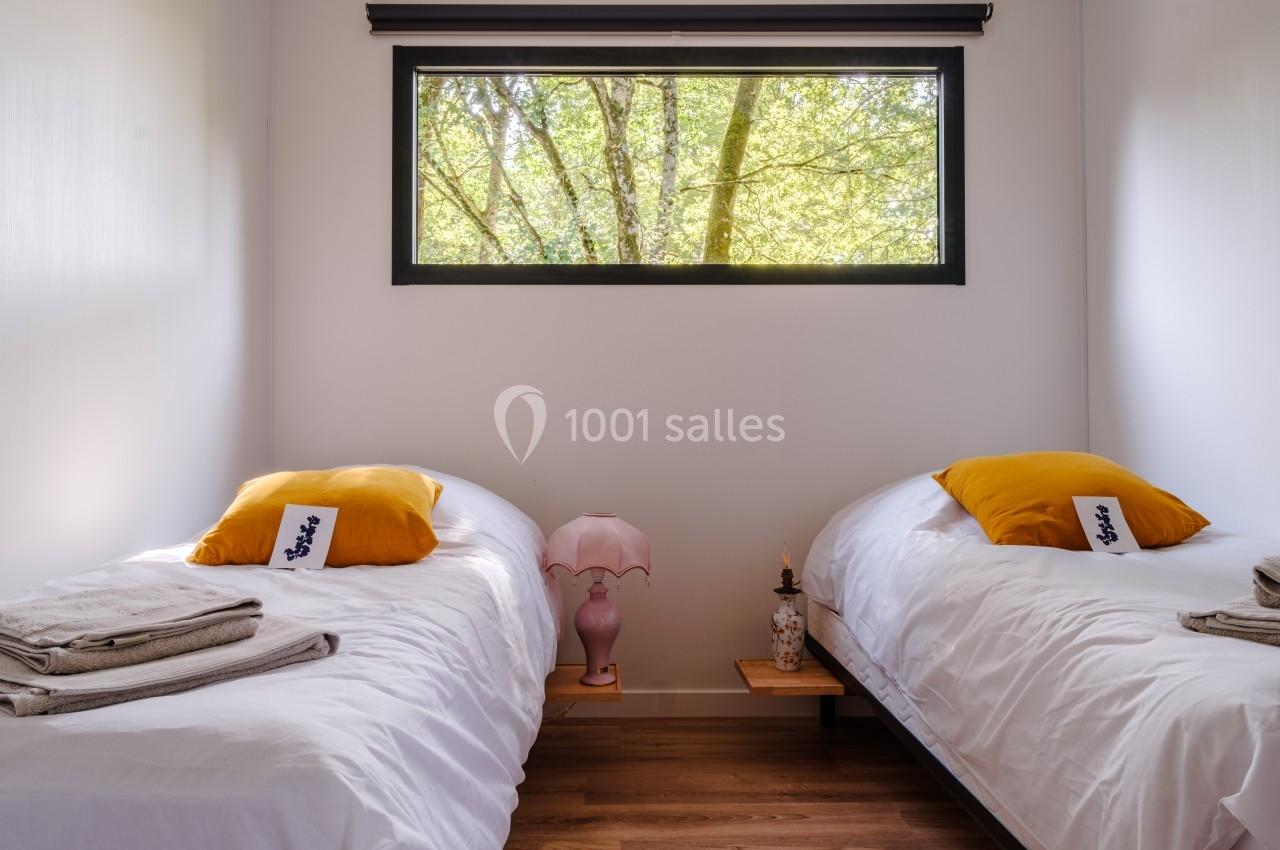 Deux lits simples avec oreillers jaunes, une lampe entre eux et une fenêtre donnant sur des arbres.