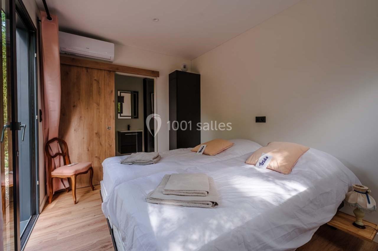 Chambre lumineuse avec deux lits simples, parquet en bois, armoire, chaise et salle de bain attenante visible.