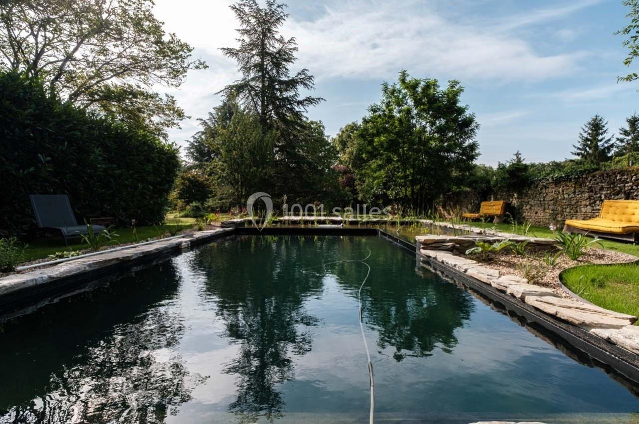 Étang de baignade entouré de végétation, avec des chaises longues et un ciel dégagé en arrière-plan.