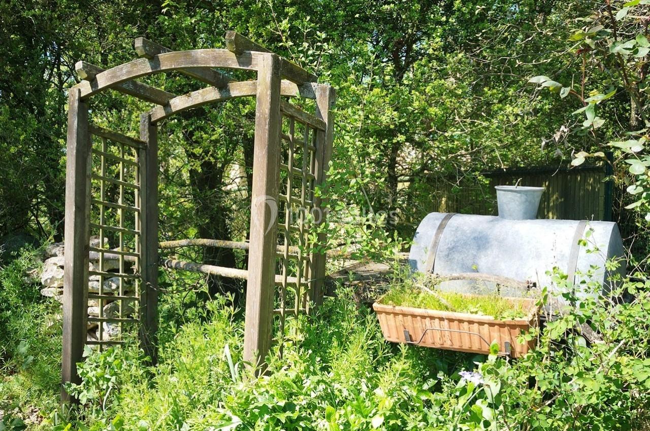 Pergola en bois entourée de végétation dense, avec une jardinière et un réservoir métallique à l'arrière-plan.