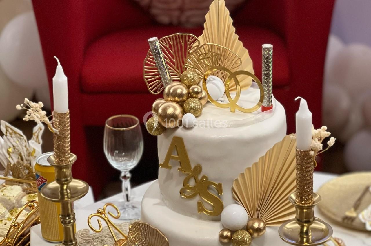 Gâteau blanc décoré de détails dorés, bougies et initiales, posé sur une table avec chandeliers et décor festif.