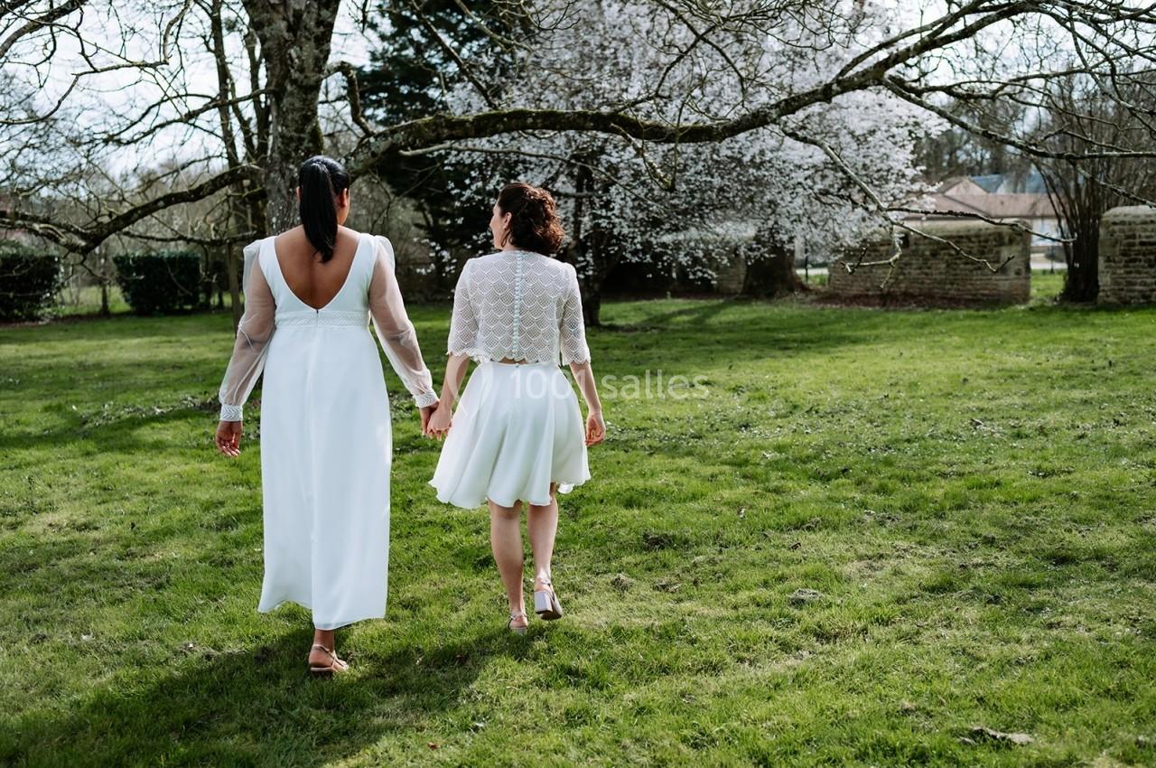 Deux femmes en robes blanches marchant main dans la main dans un jardin verdoyant avec des arbres en fleurs.