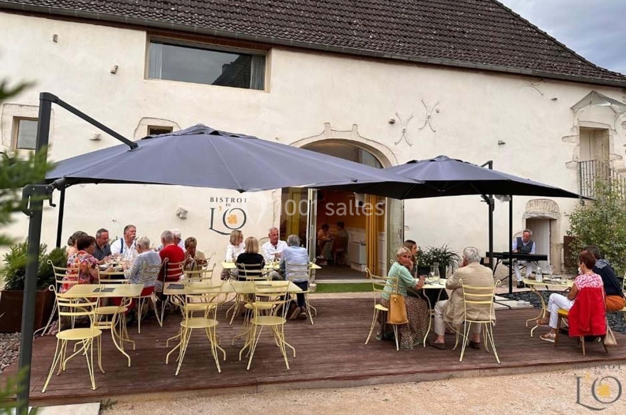 Terrasse d'un bistrot avec des clients attablés sous des parasols, devant un bâtiment ancien en pierre.