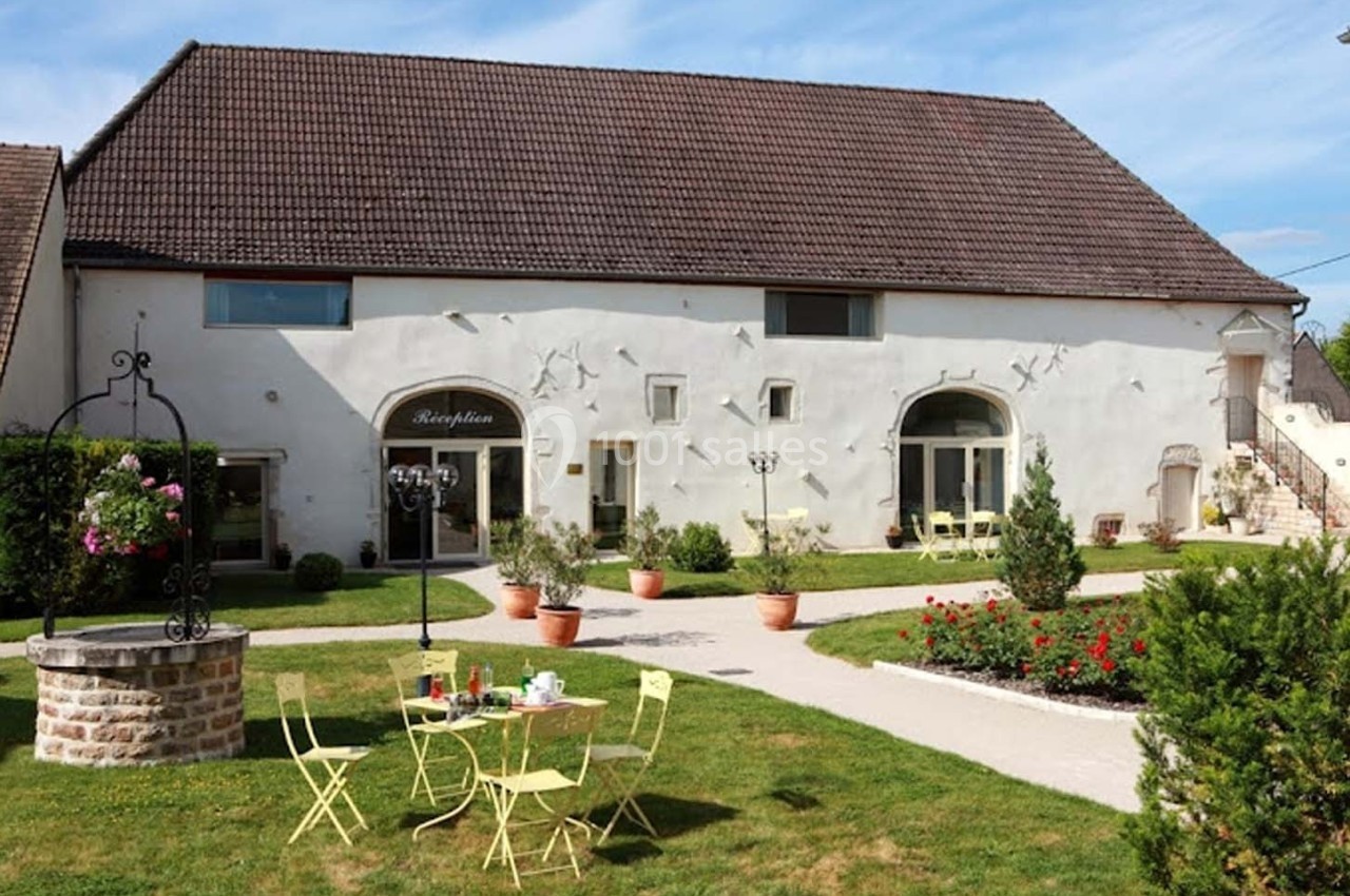 Façade d'un bâtiment ancien avec un toit en tuiles, un jardin aménagé, une table et des chaises jaunes en premier plan.