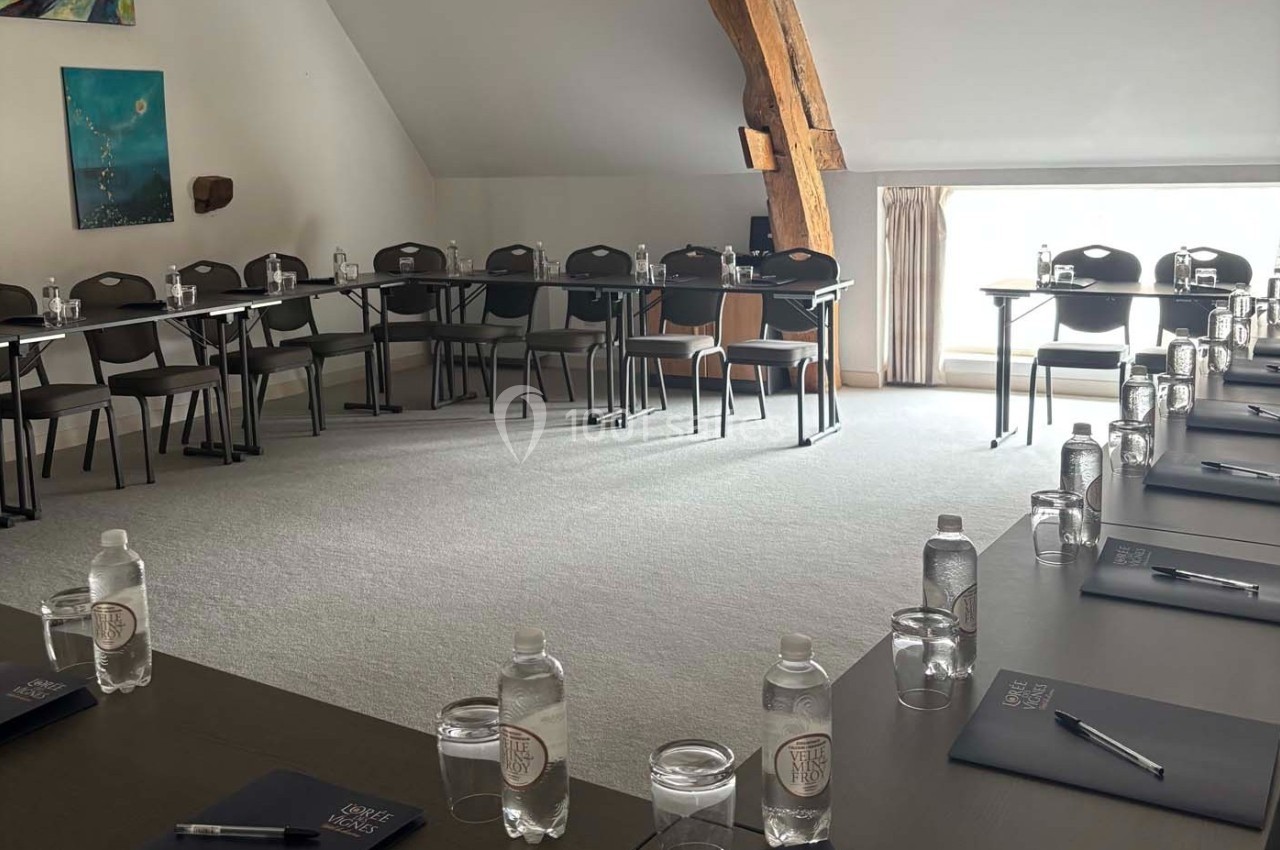 Salle de réunion lumineuse avec tables disposées en U, chaises, bouteilles d'eau, carnets et stylos.