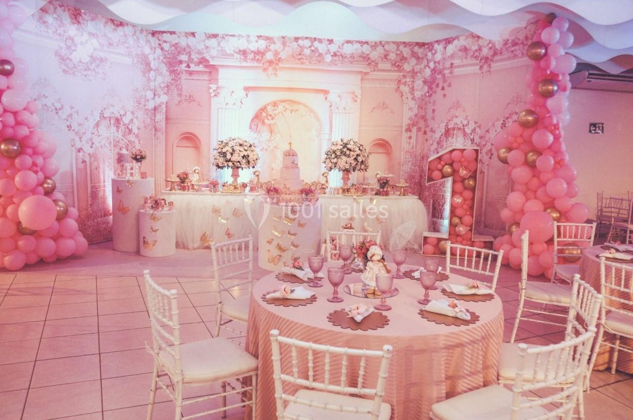 Salle décorée pour une fête d'anniversaire avec des ballons roses, une grande table centrale et des chaises blanches.