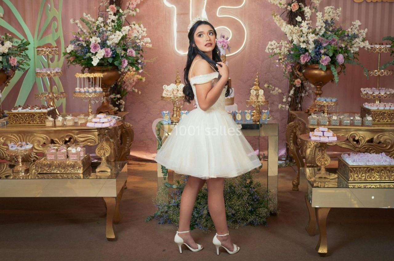 Une jeune femme en robe blanche pose devant une table décorée de fleurs et de desserts dans un cadre festif.
