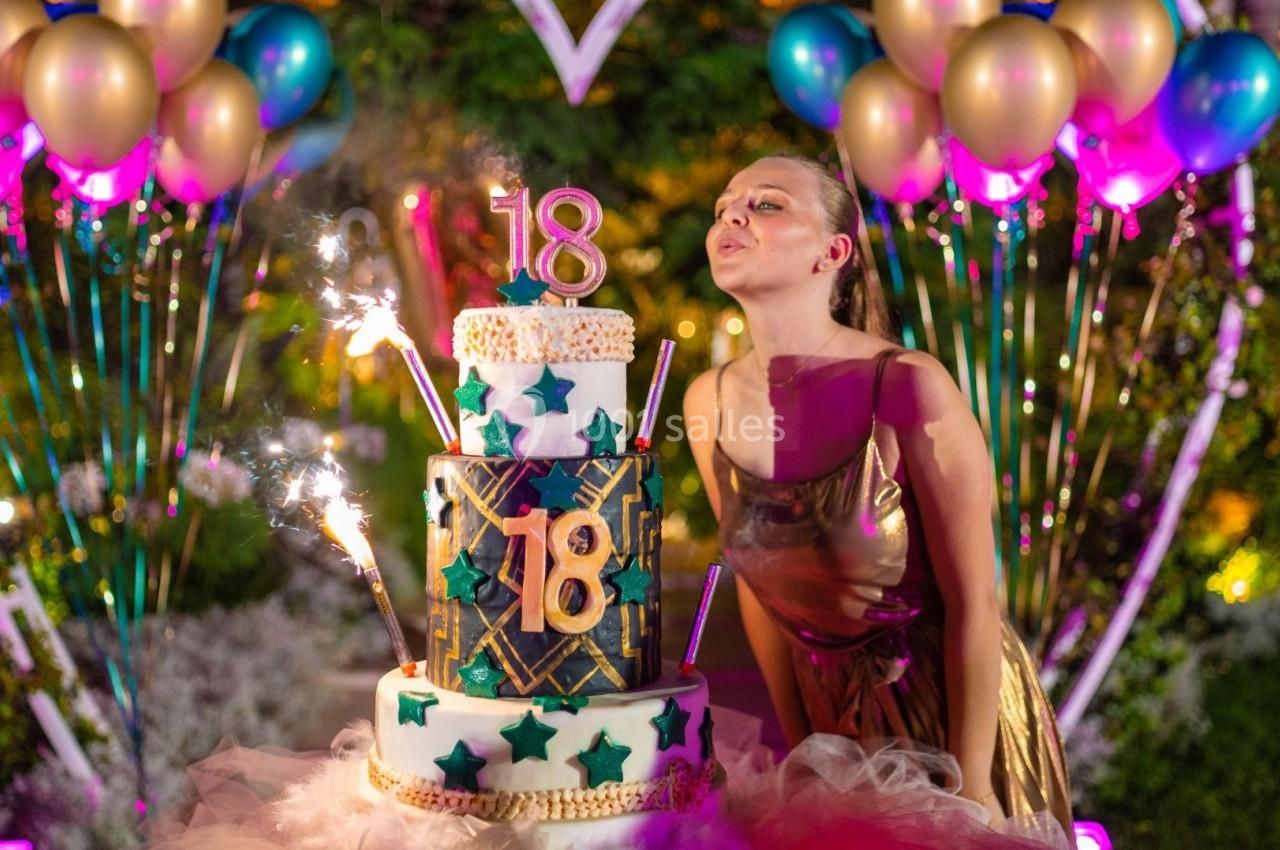 Jeune femme soufflant des bougies sur un gâteau décoré pour un 18e anniversaire, entourée de ballons colorés.