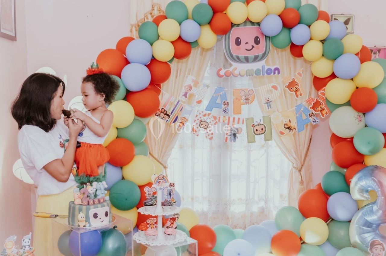 Une femme tenant un enfant devant une arche de ballons colorés et une décoration sur le thème d'anniversaire.
