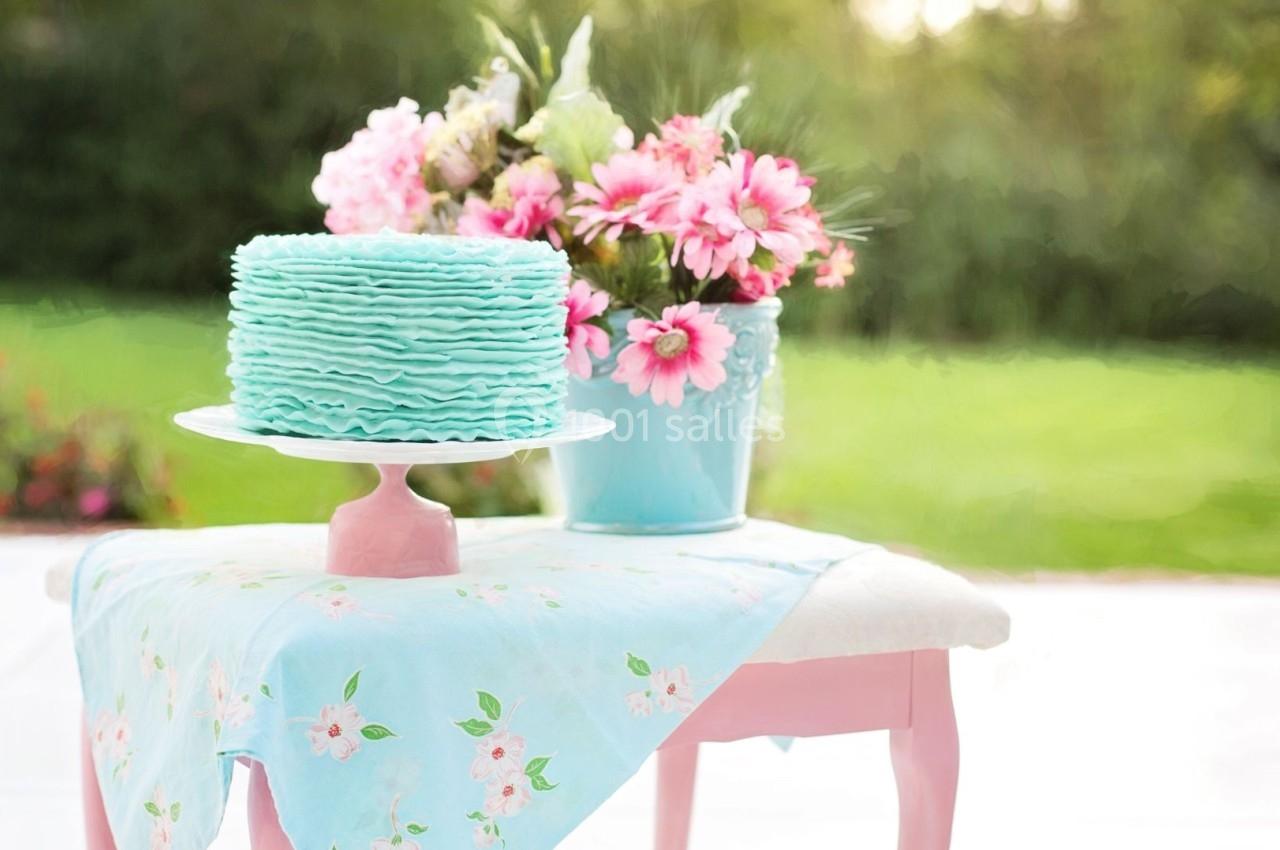 Gâteau bleu pastel sur un présentoir, posé sur une table avec une nappe fleurie, fleurs roses en arrière-plan.