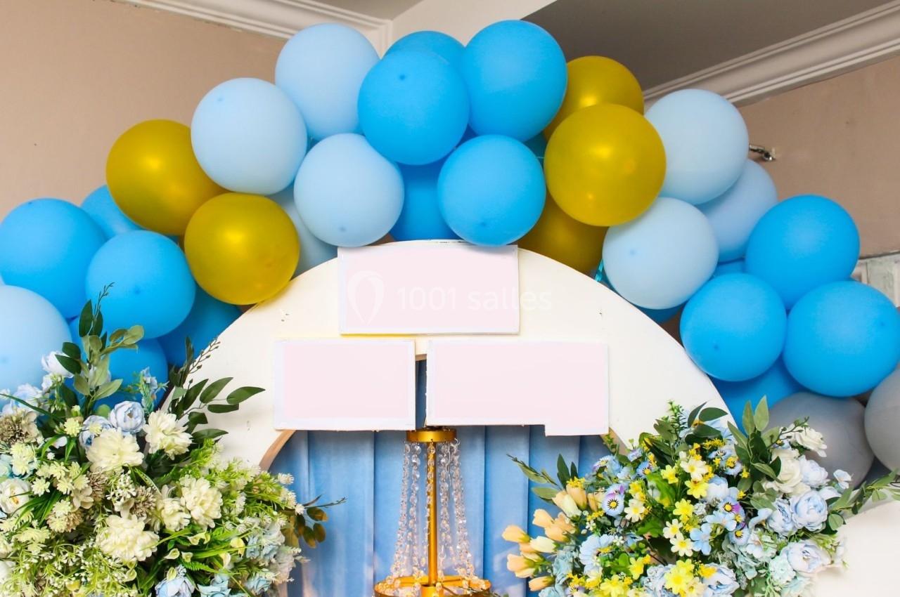 Décoration festive avec arche de ballons bleus et dorés, entourée de bouquets de fleurs colorées.