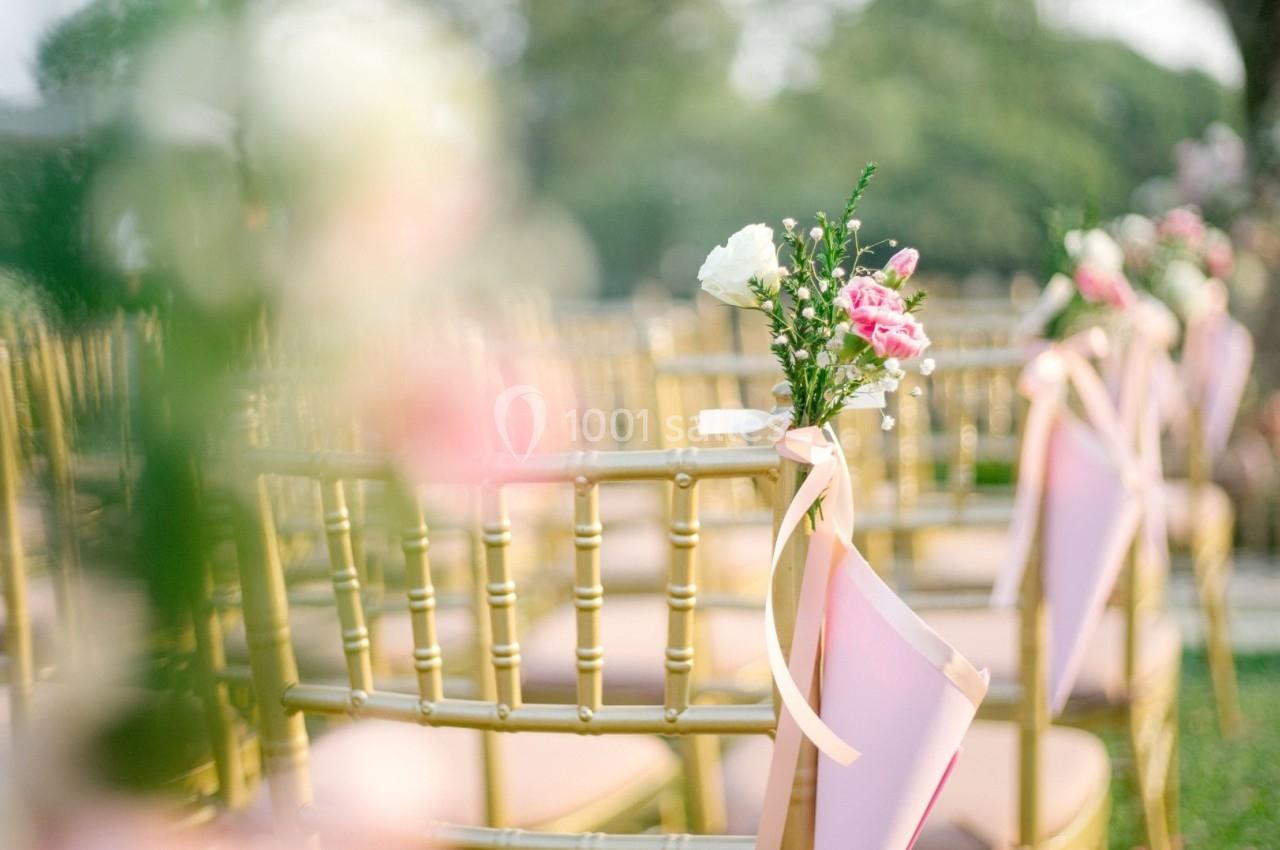 Chaises dorées décorées de rubans roses et de fleurs, alignées pour une cérémonie en plein air.